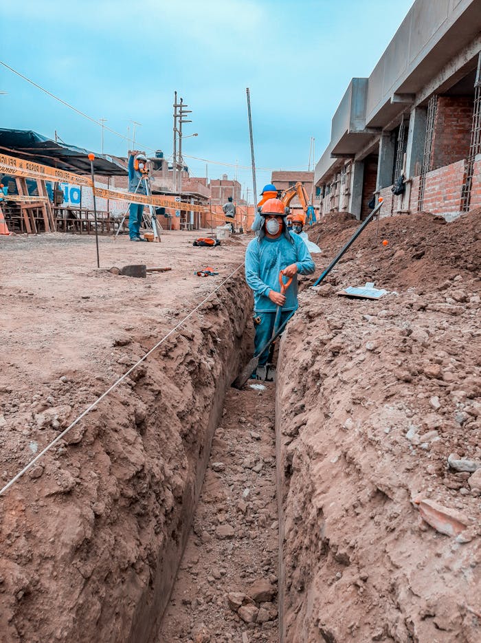 Construction workers in protective gear digging a trench at a site with buildings in the background.
